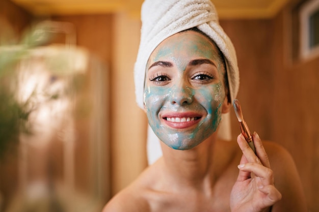 Молодая улыбающаяся женщина наносит маску для лица кистью в ванной комнате