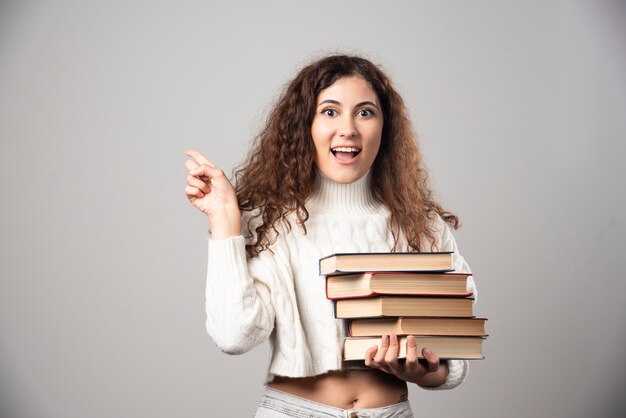 Young smiling woman carrying a lot of books on a gray wall. High quality photo