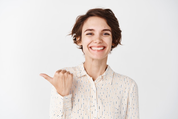 Young smiling woman employee looking happy and proud, pointing finger left company, standing against white wall