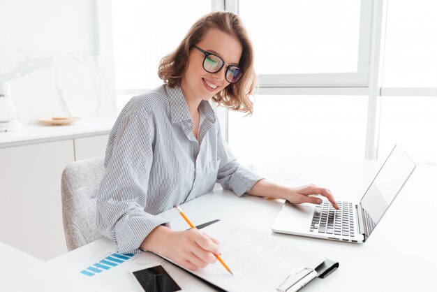 メガネとストライプのシャツの若い笑顔の女性がライトキッチンのテーブルに座りながらドキュメントとコンピューターを操作