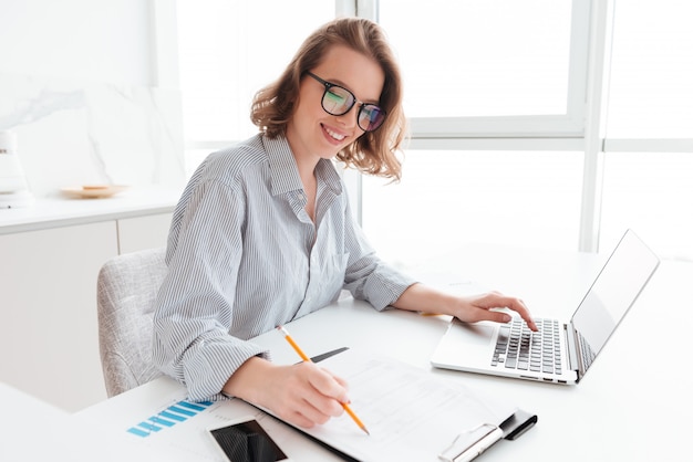 Молодая женщина улыбается в очках и полосатой рубашке, работа с документами и компьютером во время размещения за столом в светлой кухне