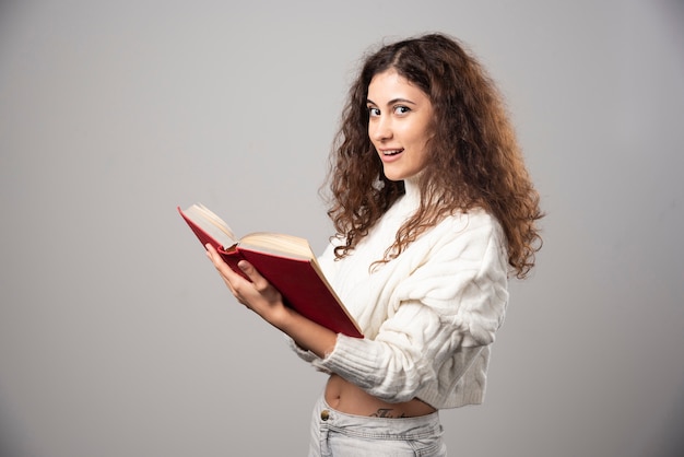 灰色の壁に赤い本を持っている若い笑顔の女性。高品質の写真