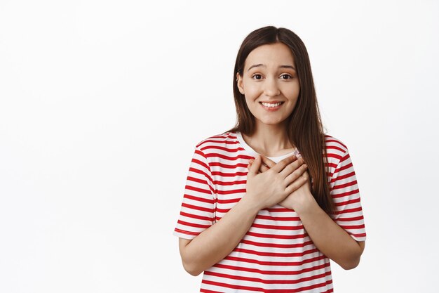 Young smiling woman looks thankful, holds hands on heart and thanking you, feel grateful, express gratitude and joy, standing against white wall