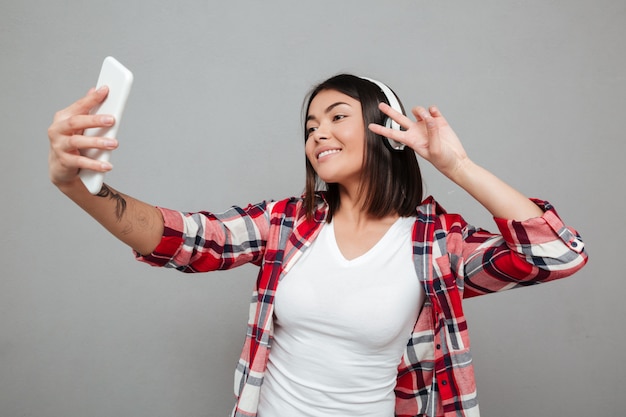 젊은 웃는 여자는 회색 벽에 selfie를 확인합니다.