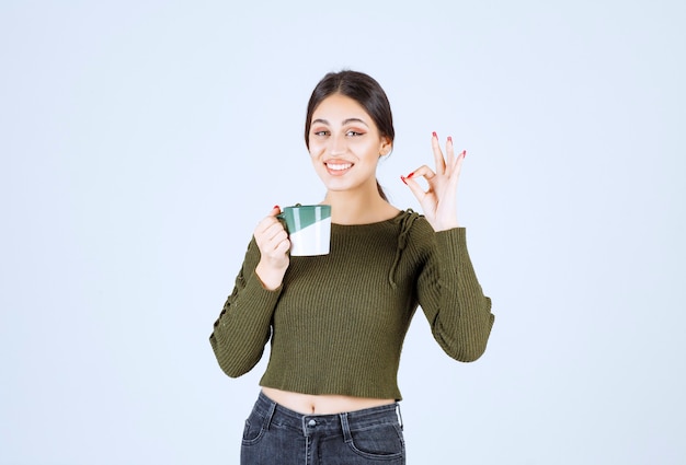 Un modello di giovane donna sorridente che tiene una tazza e che mostra gesto ok.