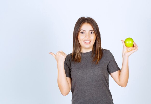 young smiling woman model holding a green apple and showing a thumb aside .