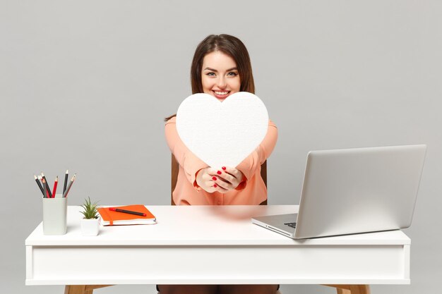 Young smiling woman in pastel casual clothes holding white heart with copy space sit, work at desk with pc laptop isolated on gray background. achievement business career lifestyle concept. mock up.