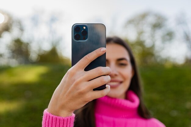 Young smiling woman in pink sweater walking in green park using phone