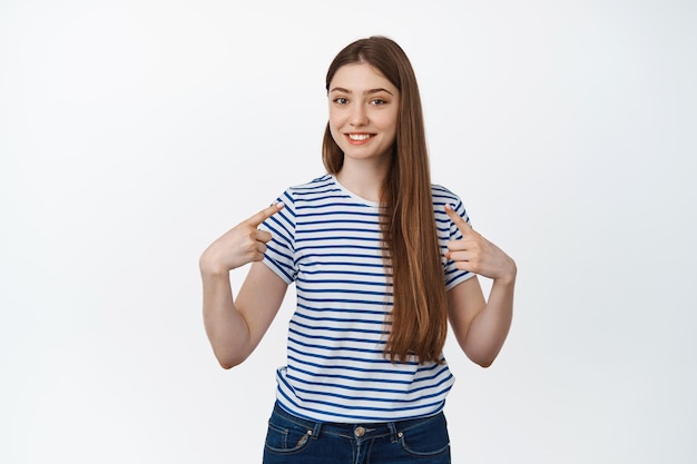 Молодая улыбающаяся женщина, указывающая пальцем на себя, самореклама, стоящая в повседневной полосатой футболке на белом фоне