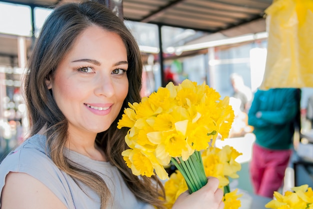 新鮮な花を選ぶ若い笑顔の女性。屋外の晴れた日に新鮮な花市場の屋台に立っている間に楽しんで花の花束を嗅ぐ美しい若い女性のプロフィールの肖像画を閉じます。