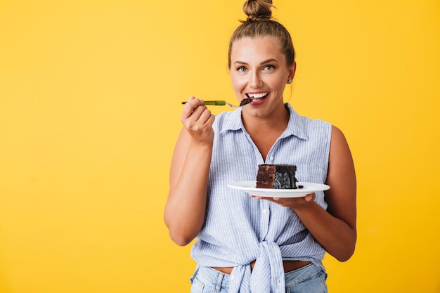 黄色の背景にチョコレートケーキを食べながらカメラで幸せそうに見てシャツの若い笑顔の女性