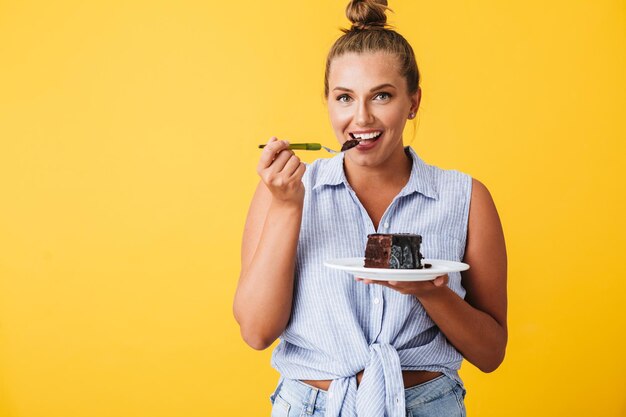 Молодая улыбающаяся женщина в рубашке счастливо смотрит в камеру, поедая шоколадный торт на желтом фоне