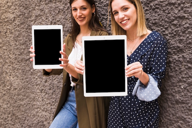 Free photo young smiling woman showing digital tablet standing against wall