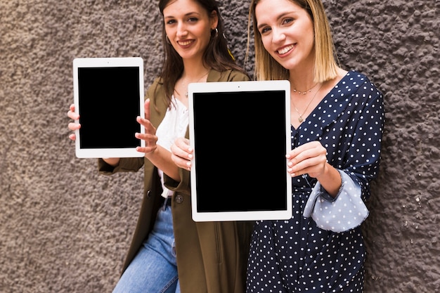Young smiling woman showing digital tablet standing against wall