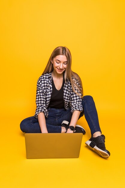 Young smiling woman sitting on floor with laptop isolated on yellow wall