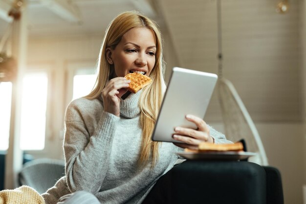 Young smiling woman surfing the net on touchpad and eating waffle while relaxing in the living room.