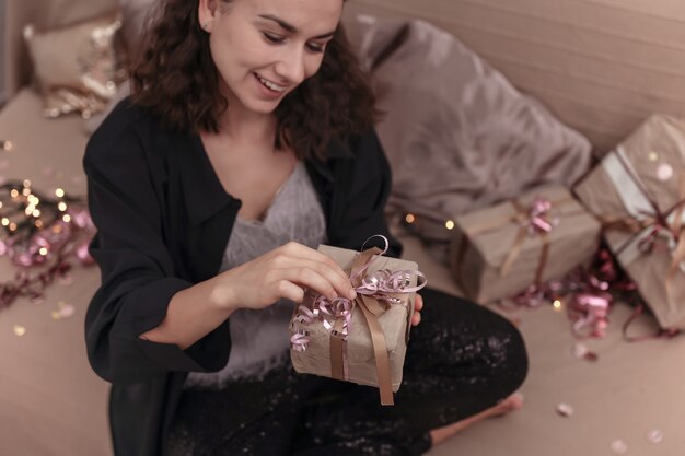 Молодая улыбающаяся женщина распаковывает рождественский подарок, сидя на кровати у себя дома.