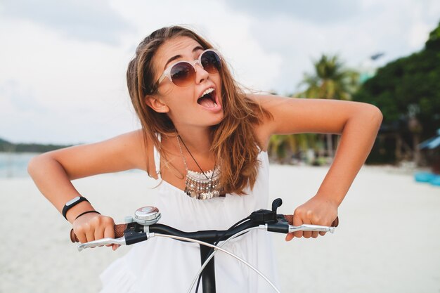 Young smiling woman in white dress riding on tropical beach on bicycle sunglasses traveling on summer vacation in Thailand