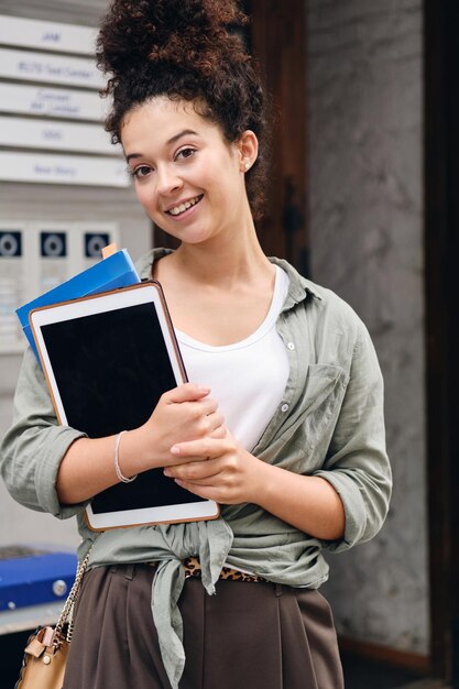 街の通りでカメラを見て喜んで教科書とタブレットを手に持っているカーキ色のシャツを着た暗い巻き毛の若い笑顔の女性