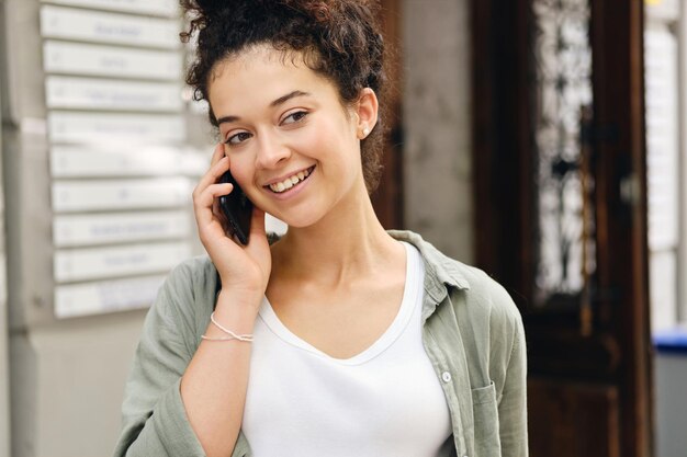 Young smiling woman with dark curly hair in khaki shirt and white Tshirt happily looking aside and talking on cellphone on city street