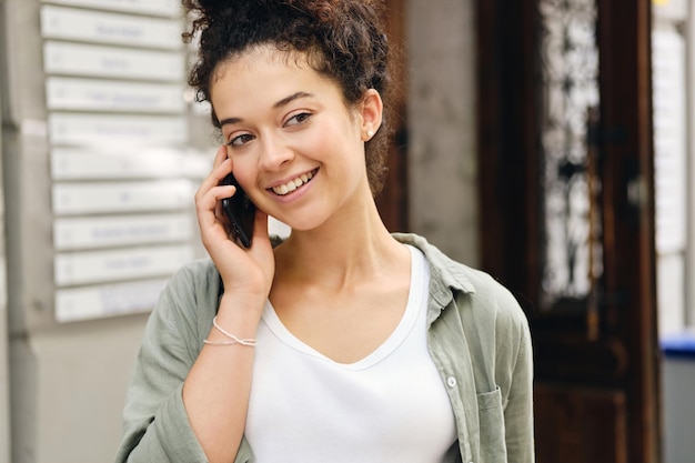 Молодая улыбающаяся женщина с темными вьющимися волосами в рубашке цвета хаки и белой футболке счастливо смотрит в сторону и разговаривает по мобильному телефону на городской улице