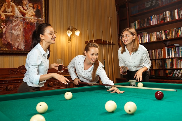 Молодые улыбающиеся женщины играют в бильярд в офисе или дома после работы.