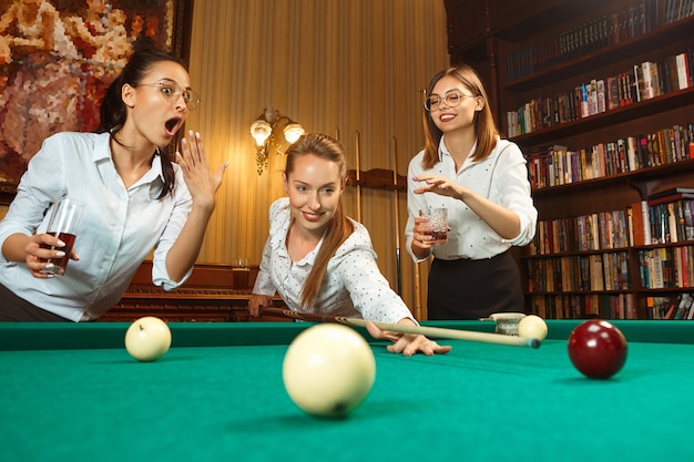 Молодые улыбающиеся женщины играют в бильярд в офисе или дома после работы.