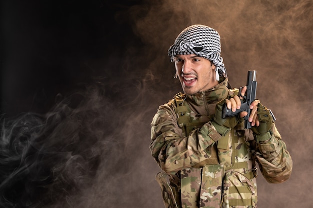 Young soldier in camouflage reloading gun on dark smoky wall