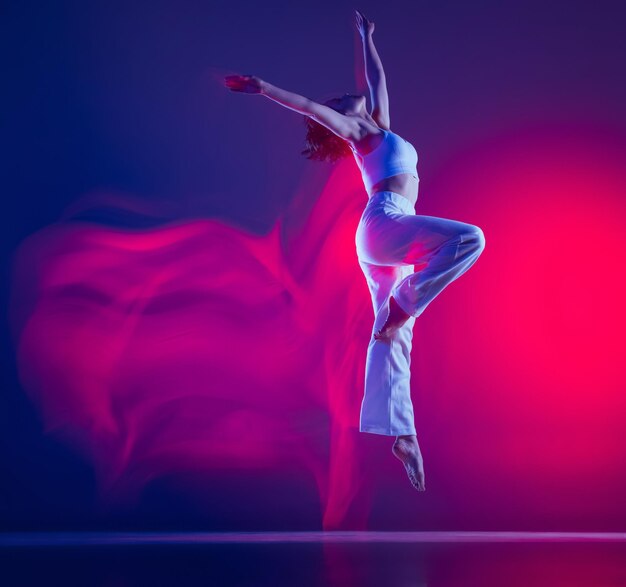 Молодая спортивная девушка в белой стильной одежде танцует хип-хоп, тренируясь на фиолетовом фоне в неоне