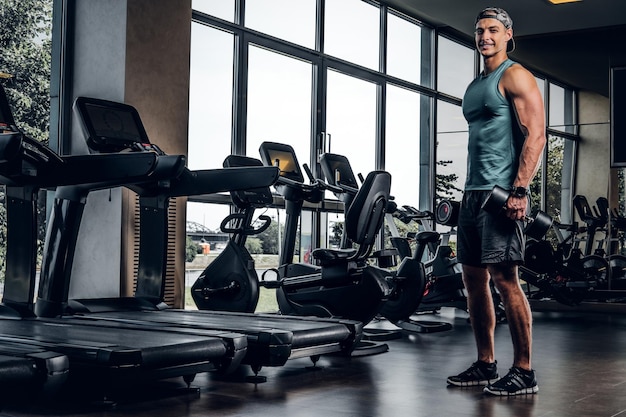 Young sportive man is doing exercises with dumbbells in empty gym club.