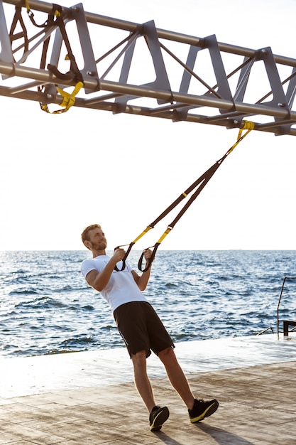 Young sportive man training with trx near sea in the morning.