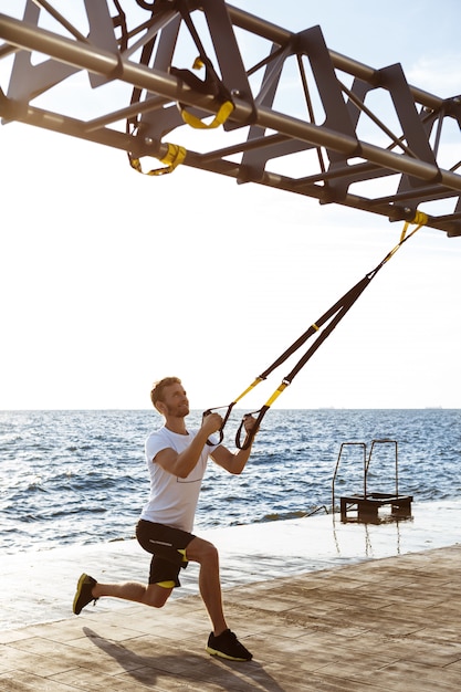 Young sportive man training with trx near sea in the morning.