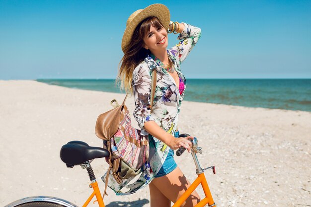 Young sportive woman in stylish white crop top and denim shorts standing on the beach with orange bicycle