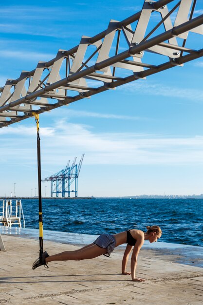 若い陽気な女性が午前中に海の近くのtrxでトレーニングします。