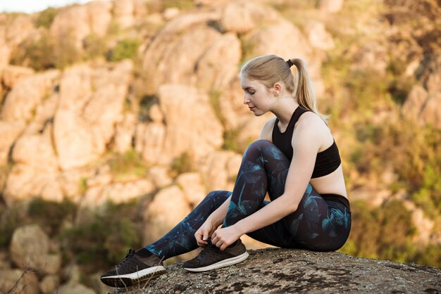 Молодая спортивная женщина завязывает шнурки, сидя на скале в каньоне