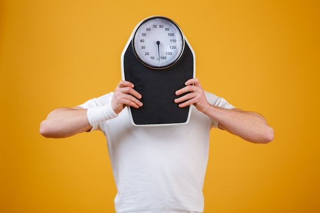 Young sports man hiding face behind weight scales