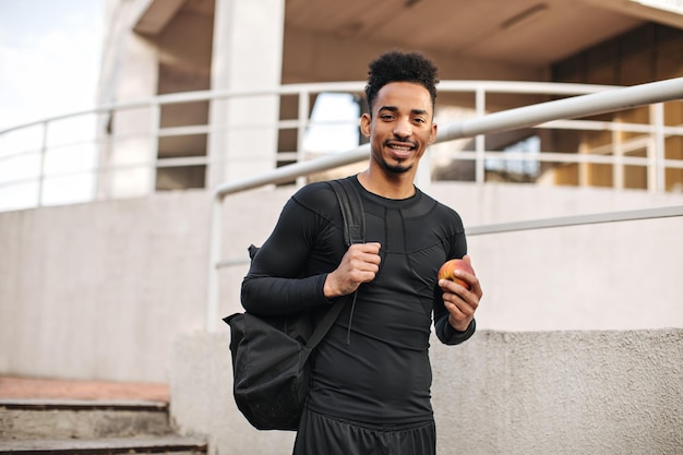 Young sportsman in black longsleeved tshirt holds backpack and fresh apple Charming guy poses outside