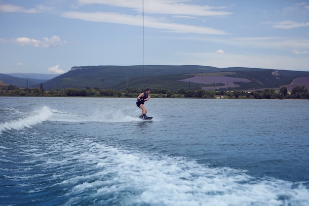 Молодой спортсмен, занимающийся серфингом через озеро. Серфер в мокром купальнике тренируется в вейк-парке, катается на вейкборде на реке, тащит моторную лодку, цепляется за кабель. Вейксерфинг, водные лыжи, спорт и отдых