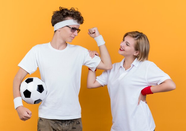 Young sporty couple man showing biceps holding soccer ball nex to his smiling girlfriend over orange