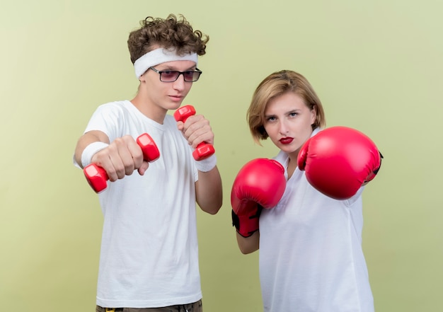 Young sporty couple man with dumbbells next to his girlfriend with boxing gloves posing over light