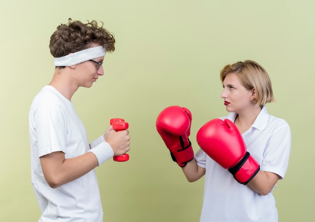 Young sporty couple man with dumbbells looking at his girlfriend with boxing gloves smiling over light