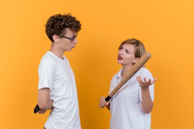 Young sporty couple man with a gun and skeptic woman with baseball bat looking at her boyfriend standing over orange wall