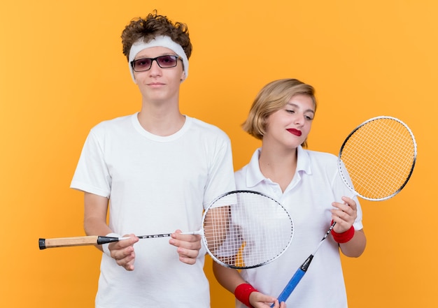 Young sporty couple man and woman holding rackets for tennis smiling confident over orange