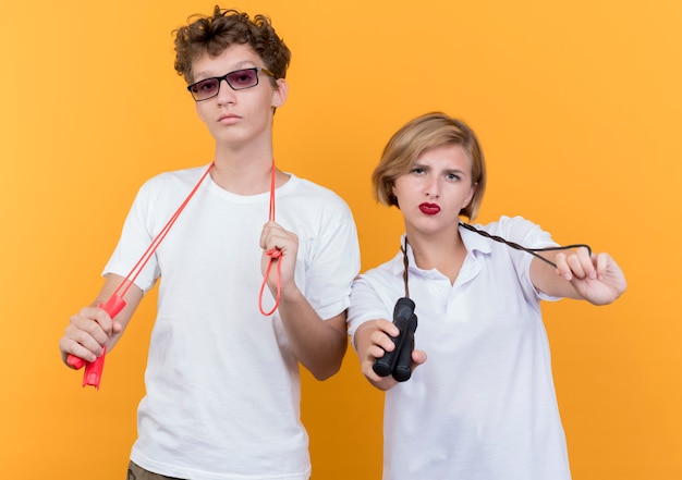 Young sporty couple man and woman holding skipping ropes with serious faces over orange