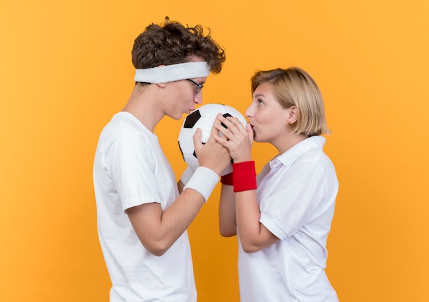 Young sporty couple man and woman looking at each other holding soccer ball and kissing it standing over orange wall