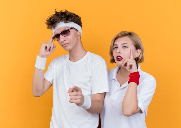 Young sporty couple man and woman  pointing with fingers at temples concentrating on a task hard standing over orange wall
