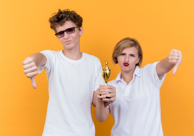 Молодая спортивная пара мужчина и женщина, стоящие вместе, держа трофей, показывая пальцы вниз, стоя над оранжевой стеной