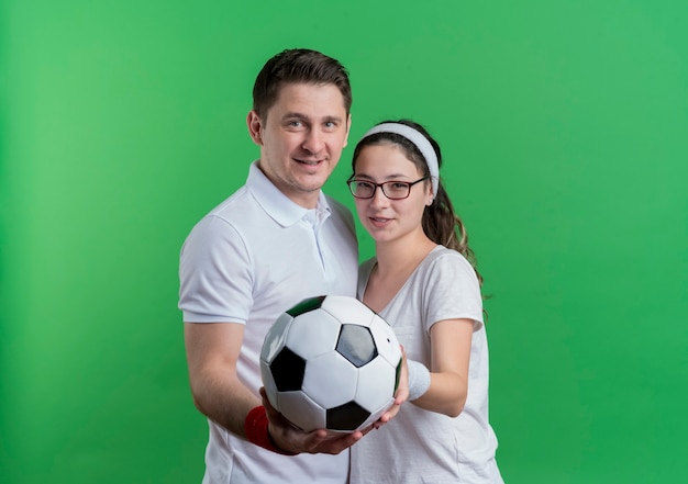 Young sporty couple man and woman together holding soccer ball smiling over green