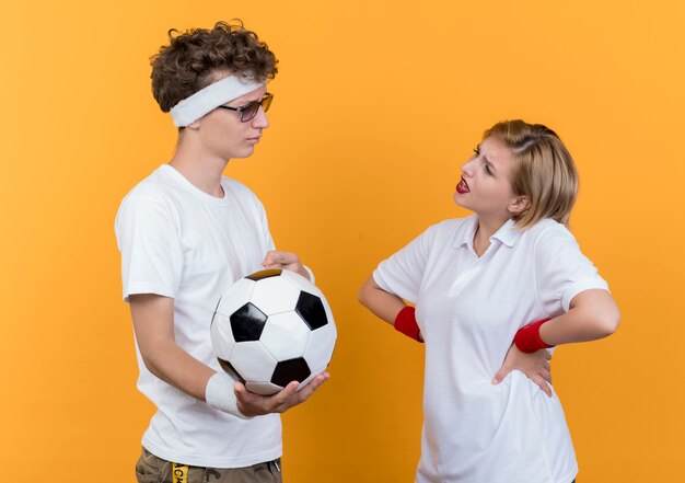 Молодая спортивная пара серьезный мужчина держит футбольный мяч, глядя на свою недовольную подругу, стоящую над оранжевой стеной