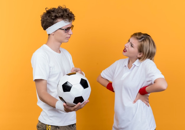 Young sporty couple serious man holding soccer ball looking at his displeased girlfriend standing over orange wall